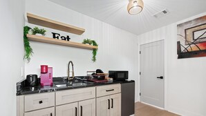 Eine moderne Küche mit schwarzem Arbeitsplatz, beigen Schränken, einer Spüle, einer Kaffeemaschine, einer Mikrowelle und einem Geschirrgestell. Darüber hängt ein Holzregal mit Pflanzen und einem Schild mit der Aufschrift 'Fat'.