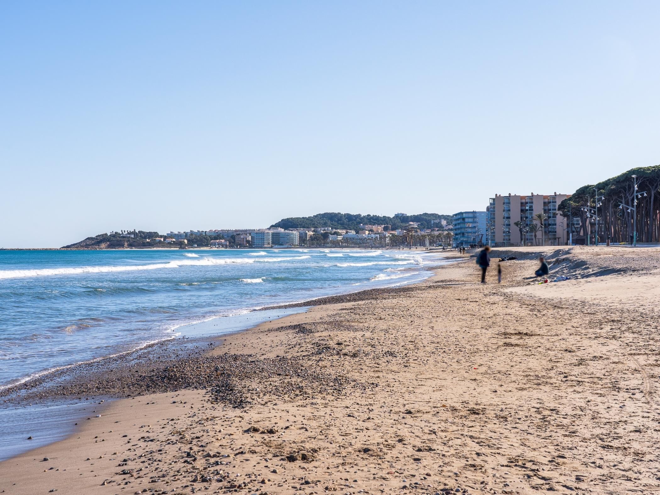 Una platja de pedres, cel serè i edificis al fons.