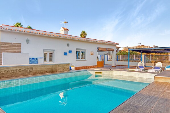 A swimming pool with a tiled deck, a covered patio area, and a building with a tiled roof and windows.