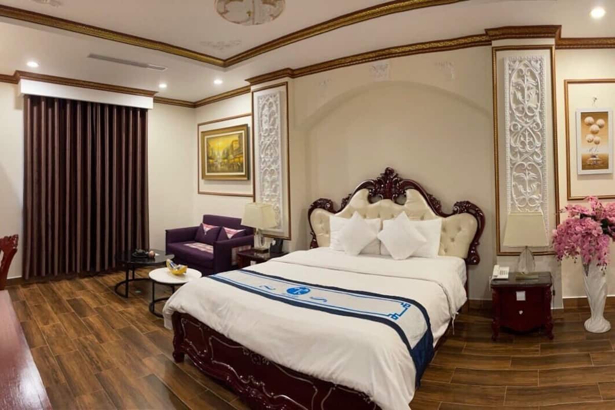 Quarto de hotel com uma cama grande, um espaço de estar com uma poltrona e mesa, um vaso com flores e um espelho na parede.