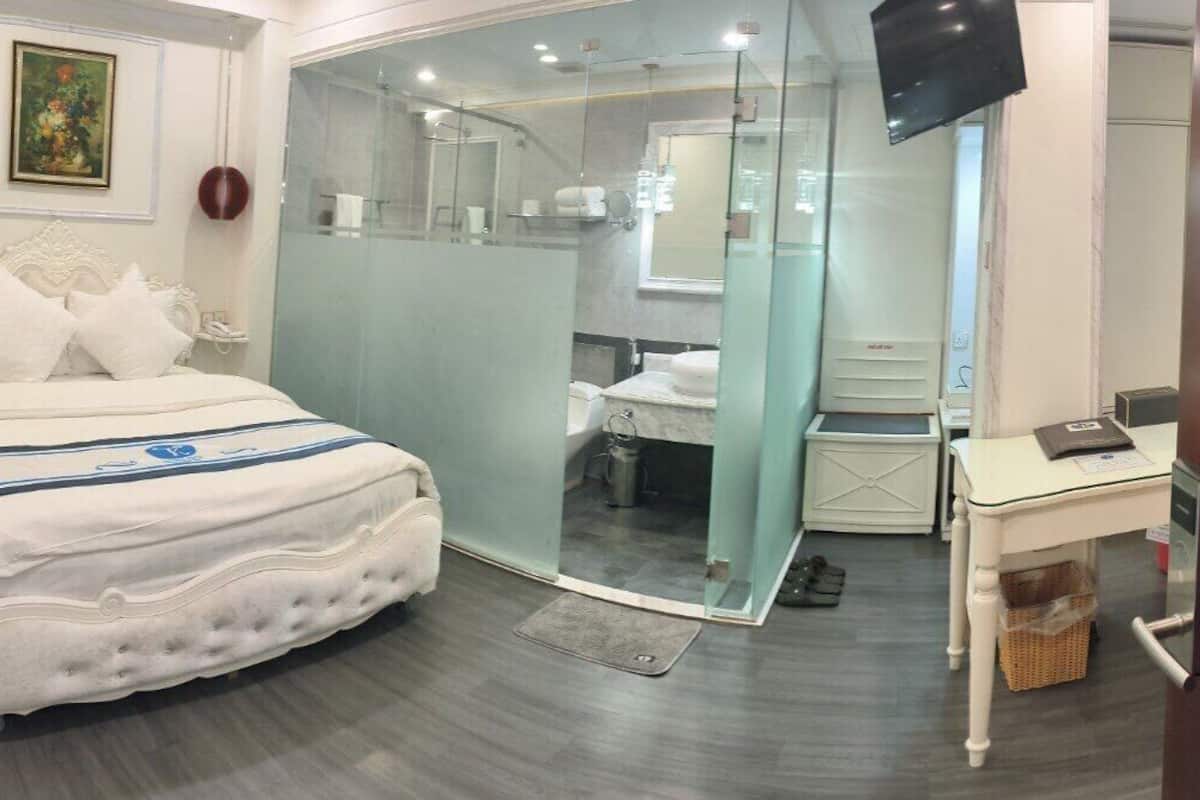 Quarto com uma cama grande, chuveiro em gabinete de vidro, uma mesinha de cabeceira e televisão fixada na parede.