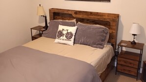 Un dormitorio con cabecera de madera, cama con manta color beige, dos almohadas, un cuadro enmarcado de un río y dos mesitas de noche con lámparas.