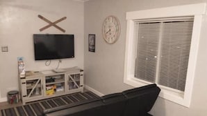 Una sala de estar con un sofá negro, un televisor montado en la pared, un reloj y una ventana con persianas.