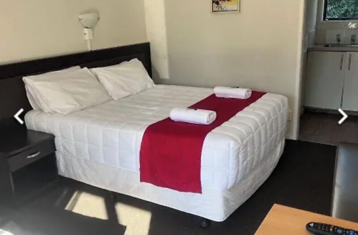 A neatly made bed with white bedding and a red blanket, a bedside table with a lamp, and a small wooden table with a remote control.