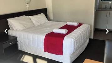 Una cama bien hecha con ropa de cama blanca y una manta roja, una mesita de noche con una lámpara, y una pequeña mesa de madera con un control remoto.