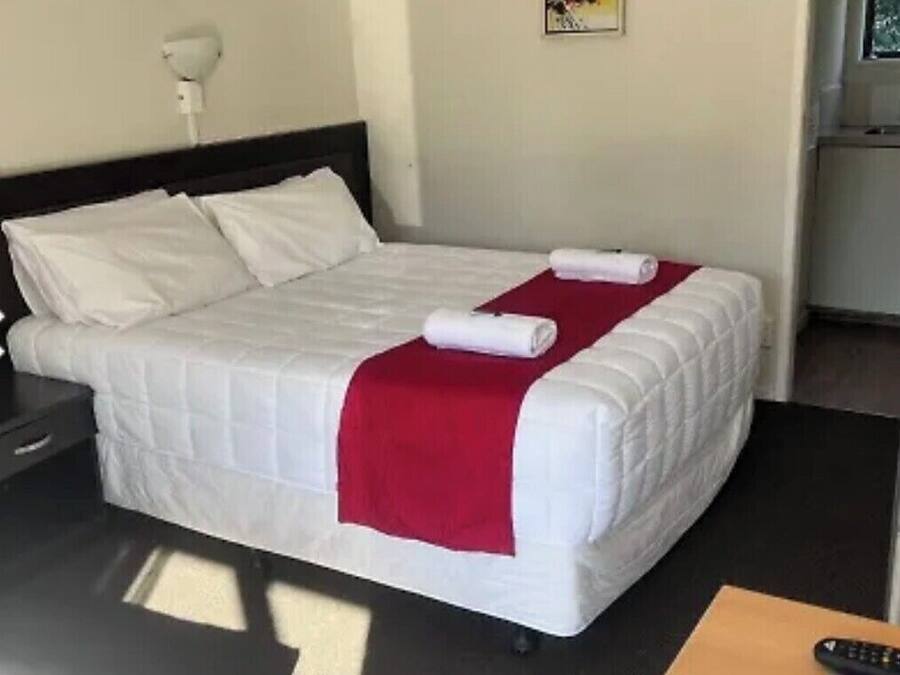 Una cama bien hecha con ropa de cama blanca y una manta roja, una mesita de noche con una lámpara, y una pequeña mesa de madera con un control remoto.