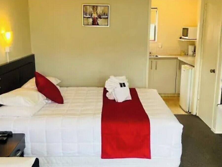 A hotel room with a bed, a red throw, a white blanket, a pillow, a bedside table, a lamp, a picture on the wall, a microwave, and a kitchenette.