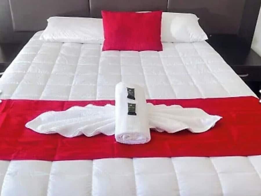 Una cama bien hecha con una colcha roja y blanca, una toalla blanca enrollada y un cojín rojo.