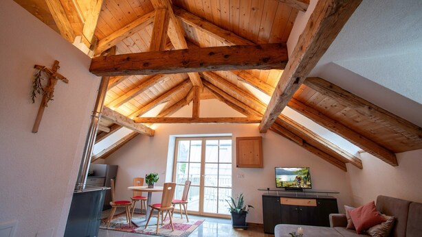 Ein Zimmer mit hohen Decken und Holzbalken, einem Essbereich mit Stühlen und Tisch, einem Fernseher und Blick durch ein großes Fenster.