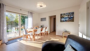 Uma sala de estar moderna com um sofá preto, um espaço de jantar com mesa de madeira e cadeiras brancas, e uma grande porta de vidro deslizante que dá para uma área externa.