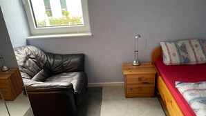 Een slaapkamer met een leren bank, een houten nachtkastje met een lamp, een houten bed met een rode sprei, en een raam met uitzicht op gebouwen en bomen.