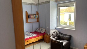 Een kleine slaapkamer met een enkel bed, een leren fauteuil, een boekenkast en een raam met uitzicht op gebouwen.