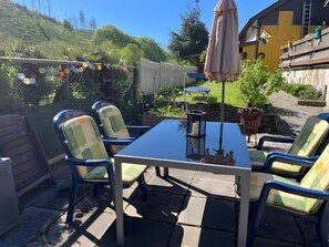 Een terras buiten met een tafel en stoelen, omringd door een houten omheining en begroeiing.