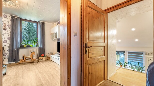 Une pièce avec une porte en bois, un radiateur, une fenêtre avec des rideaux, un cheval à bascule en bois et une vue sur l’extérieur.