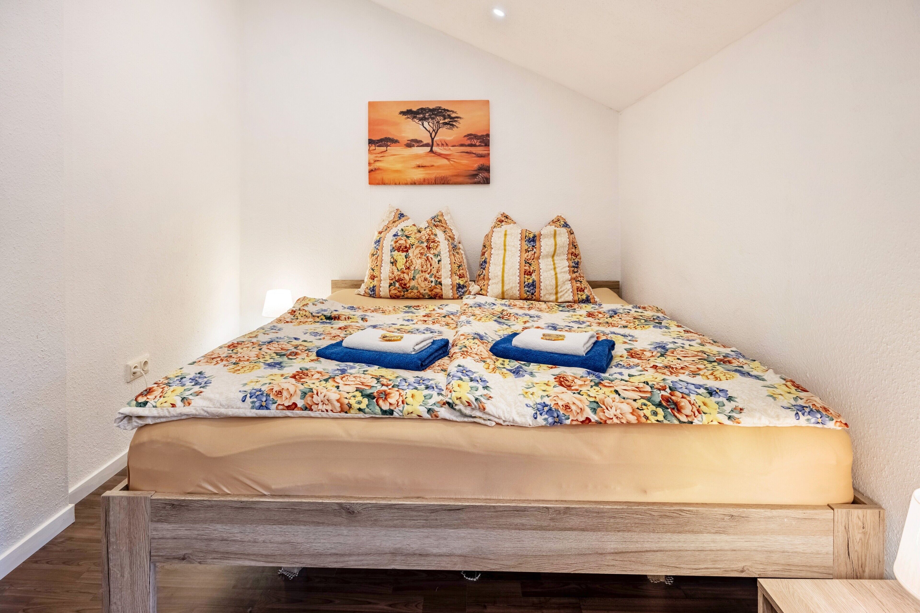 A bed with a floral bedspread, two pillows, and a painting of a tree on the wall.