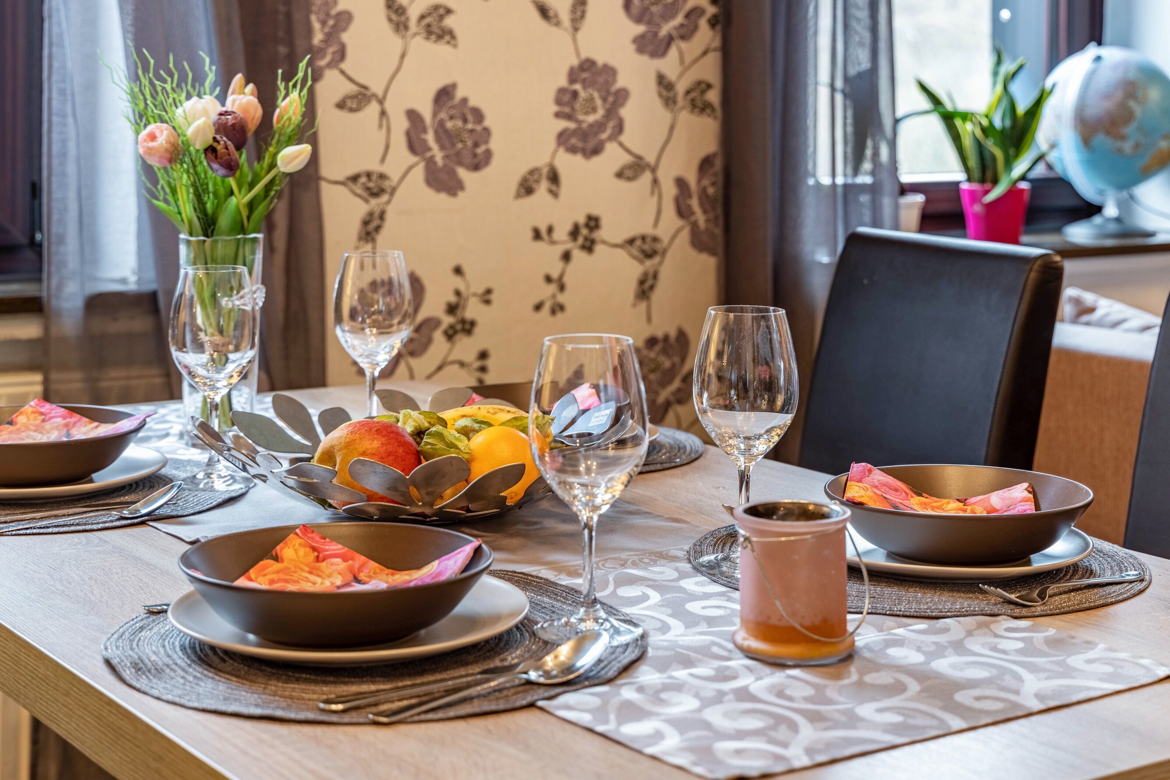 Ein Esstisch ist gedeckt mit Tellern, Schüsseln, Gläsern und Besteck. Im Hintergrund steht eine Blumenvase und ein Globus.