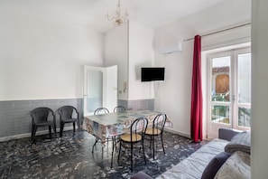 A room with a dining table, chairs, a TV, a balcony with a red curtain, and a bed.