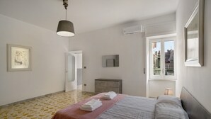 Une chambre d’hôtel avec un lit, une table de chevet, une lampe, une fenêtre donnant sur un paysage agréable, une porte et un tableau encadré au mur.