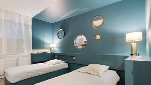Ein Hotelzimmer mit zwei Einzelbetten, einem Fenster mit durchscheinenden Vorhängen, einem Nachttisch mit Lampe und einem wandmontierten Spiegel.