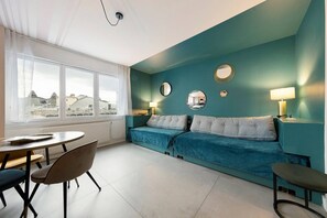Une pièce avec un canapé de couleur turquoise, une table à manger ronde entourée de chaises, et une vue sur les bâtiments à travers de grandes fenêtres.