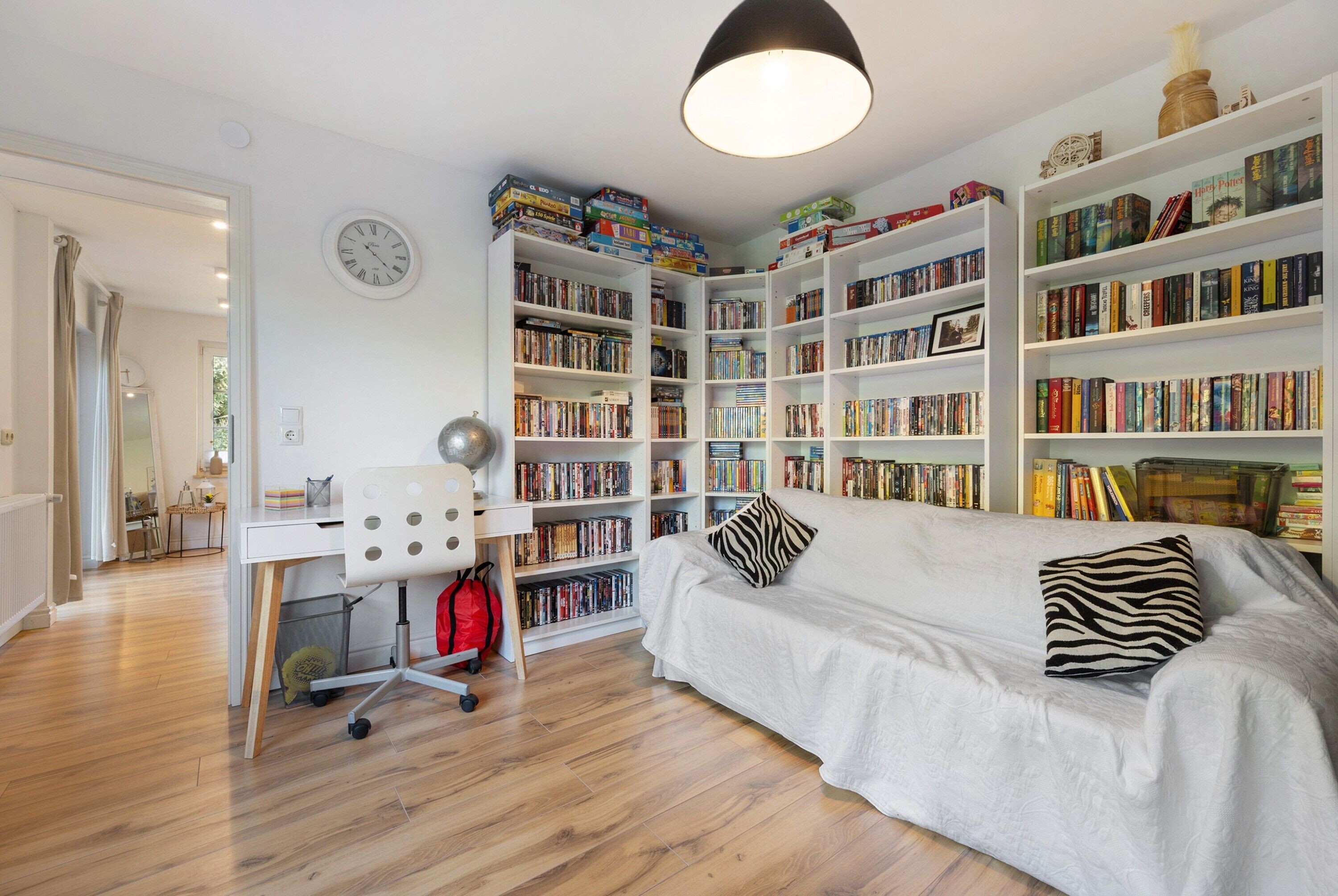Una habitación con un sofá blanco, una estantería llena de libros, un escritorio con una silla y un reloj en la pared.