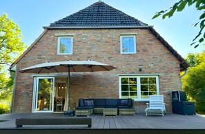 Et murstenshus med saddeltag, en terrasse med sofa og parasol, samt et fjernsyn på dækket.