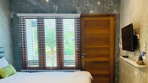 Un dormitorio con una cama, una puerta de madera, un televisor montado en la pared y una ventana con persianas.