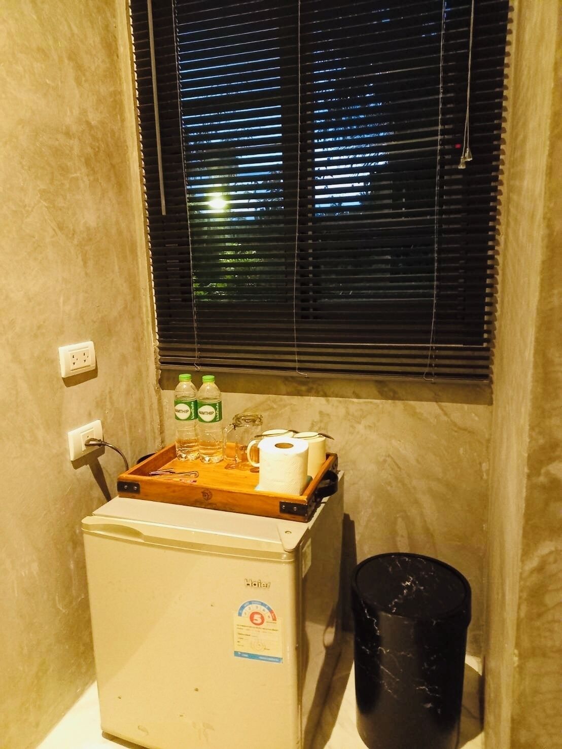 A compact room with a small refrigerator, a window with blinds, a trash bin, and a wooden tray with toiletries.