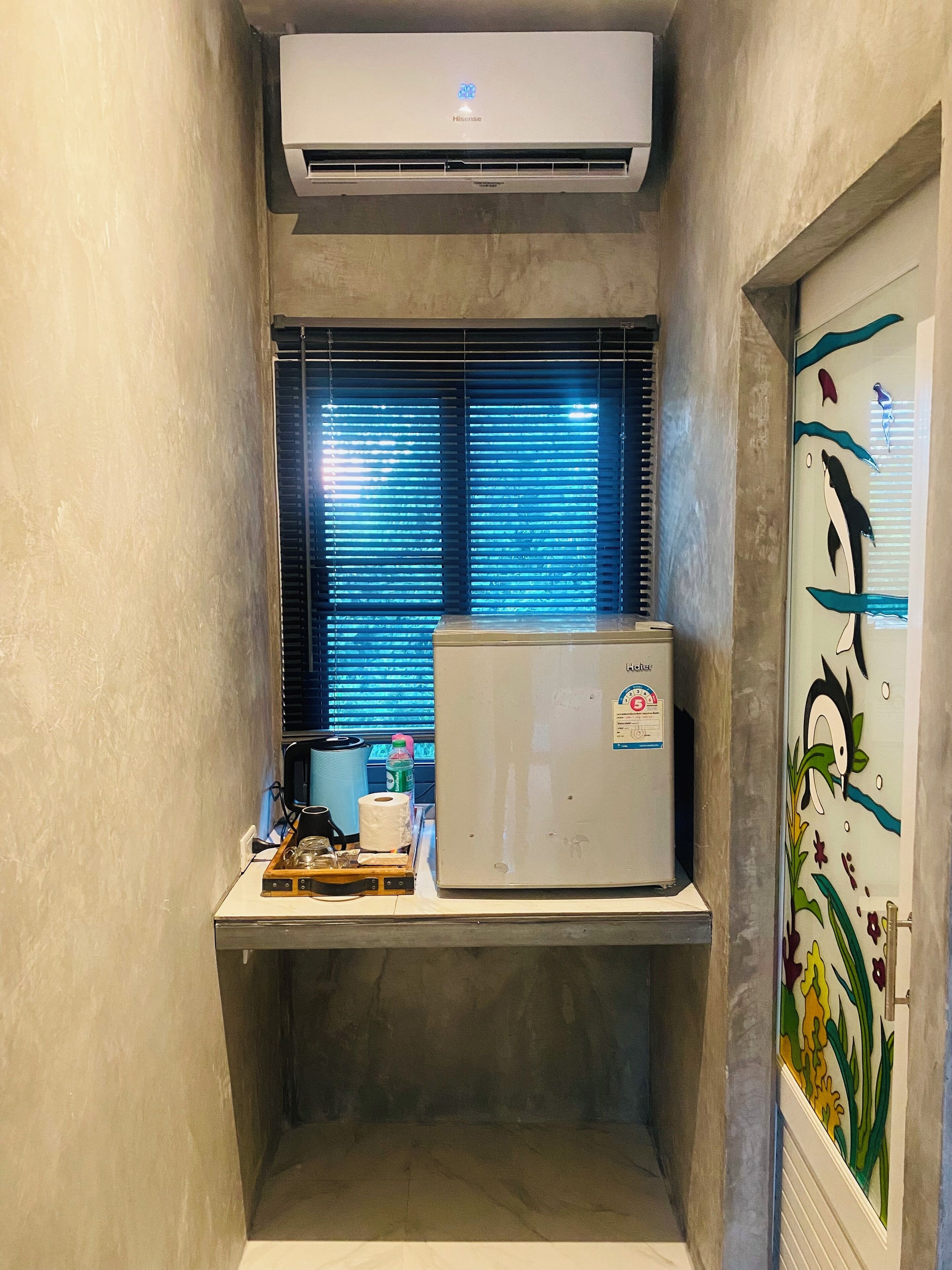 A compact room with a built-in air conditioner, a small refrigerator, and a window with blinds.