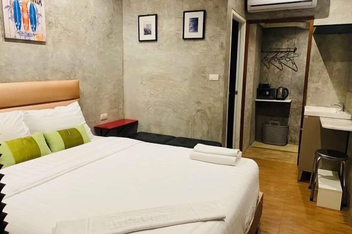 A hotel room with a bed, a desk, a chair, a small stool, a red nightstand, a wall-mounted shelf with a kettle, and a trash bin.