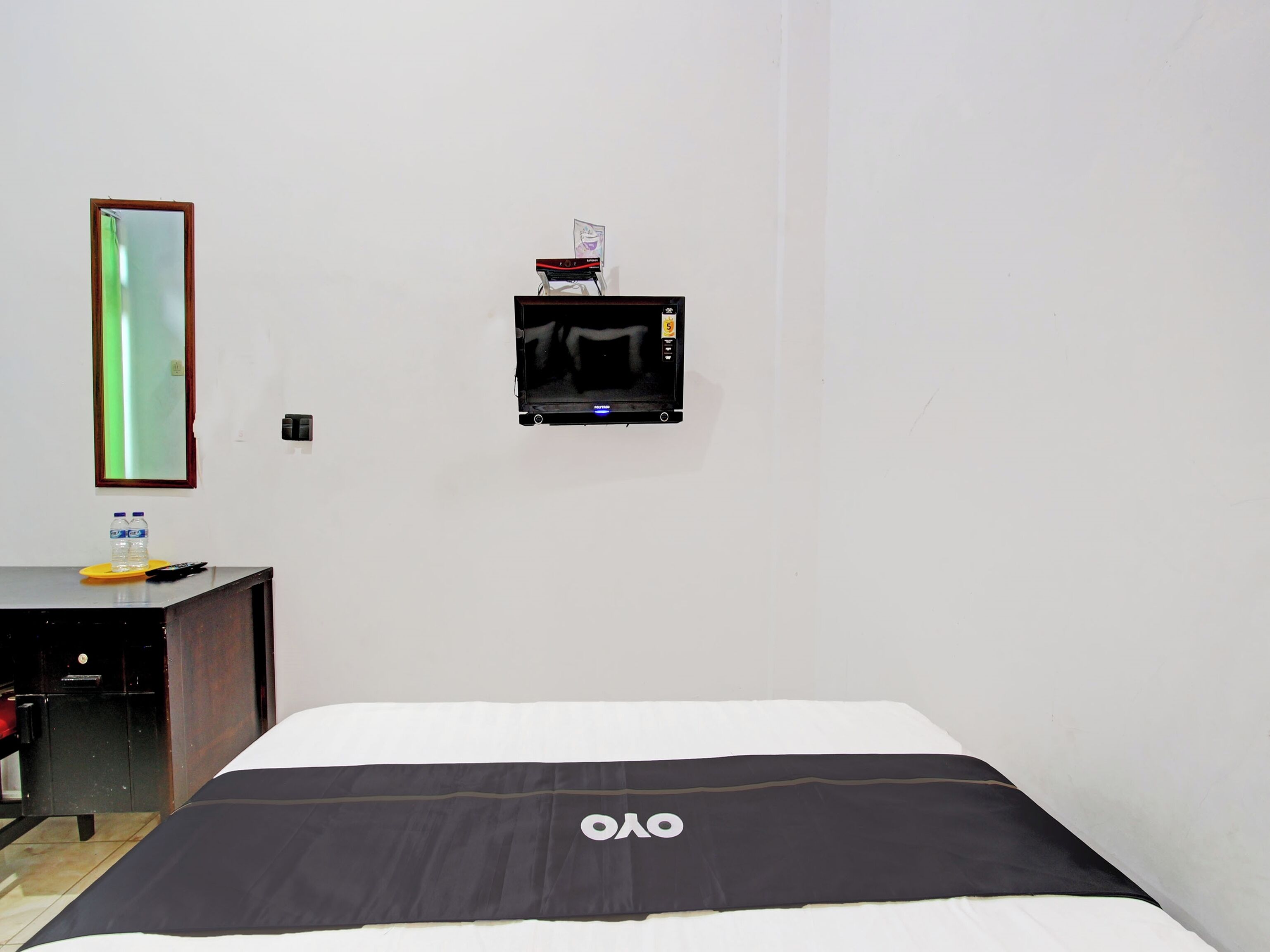 A hotel room with a bed, a small desk with a mirror, and a television on the wall.