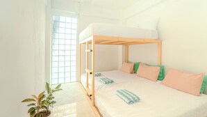 Un lit superposé avec une structure en bois, une literie blanche et des coussins verts et pêche. Une plante en pot est posée sur le sol.