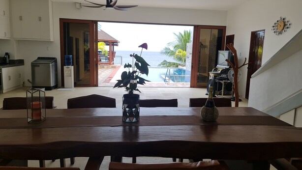 A dining area with a view of the ocean, a ceiling fan, and a table with a bottle and a plant. - Beachfront Villa with Infinity Pool, Well-Equipped Kitchen & Breathtaking Views (Batangas)