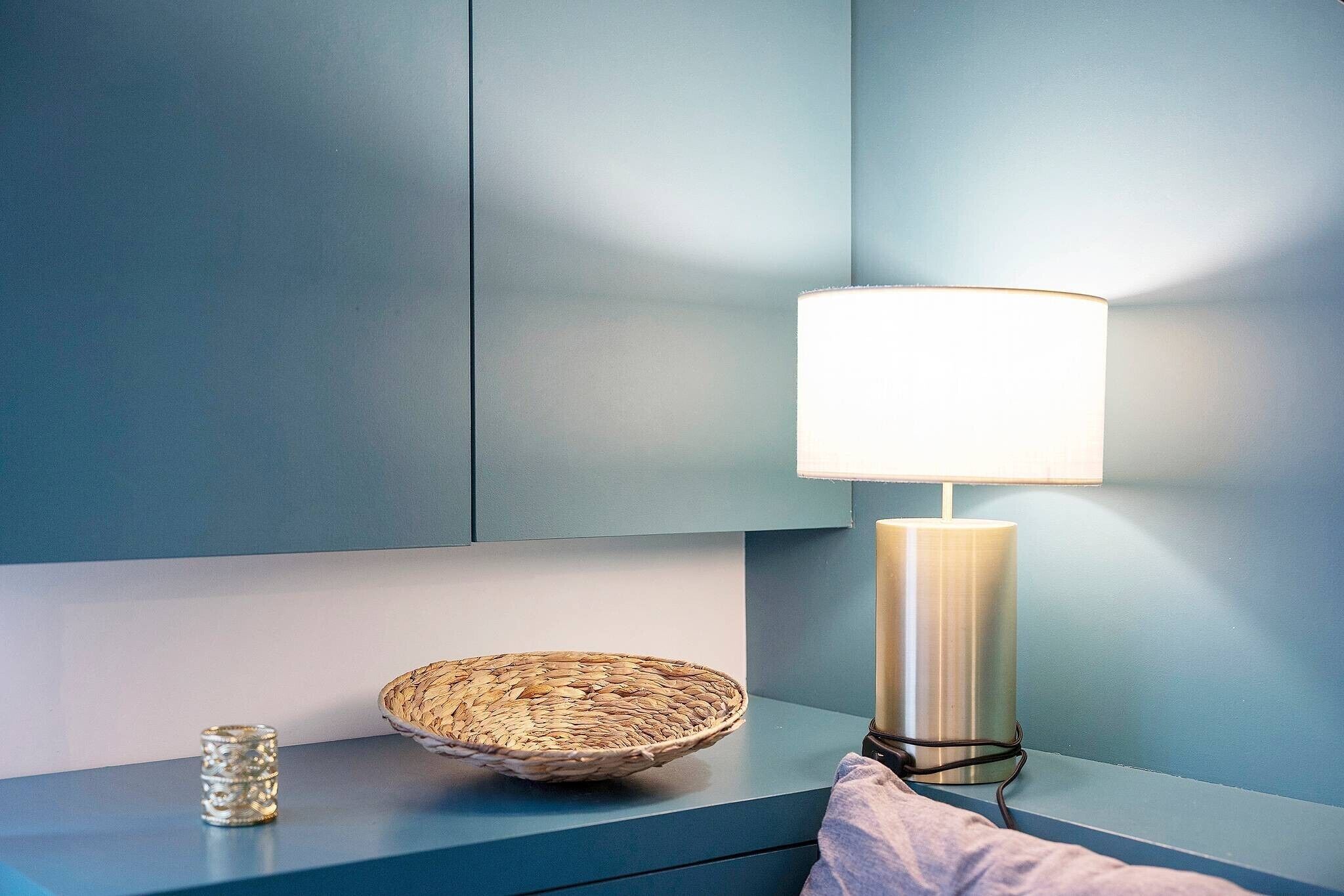 Una mesita de noche con una cesta tejida, un recipiente de vidrio y una lámpara frente a una pared azul.