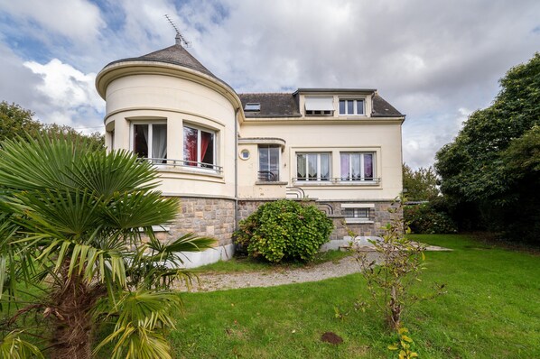Une maison à deux étages, dotée d’une tour ronde, d’un balcon et d’un jardin bien entretenu.