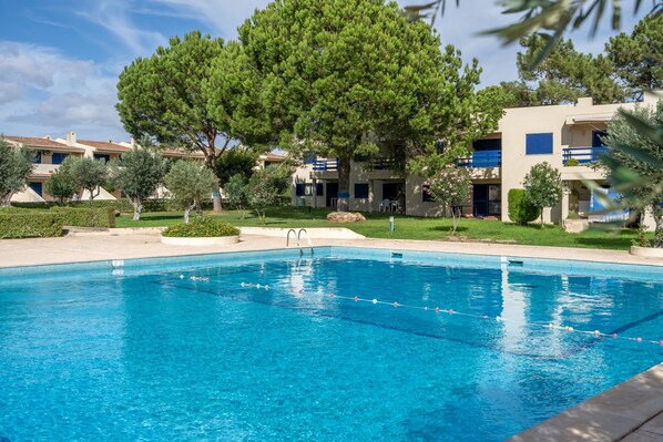 A swimming pool with clear blue water, surrounded by greenery and buildings with balconies.