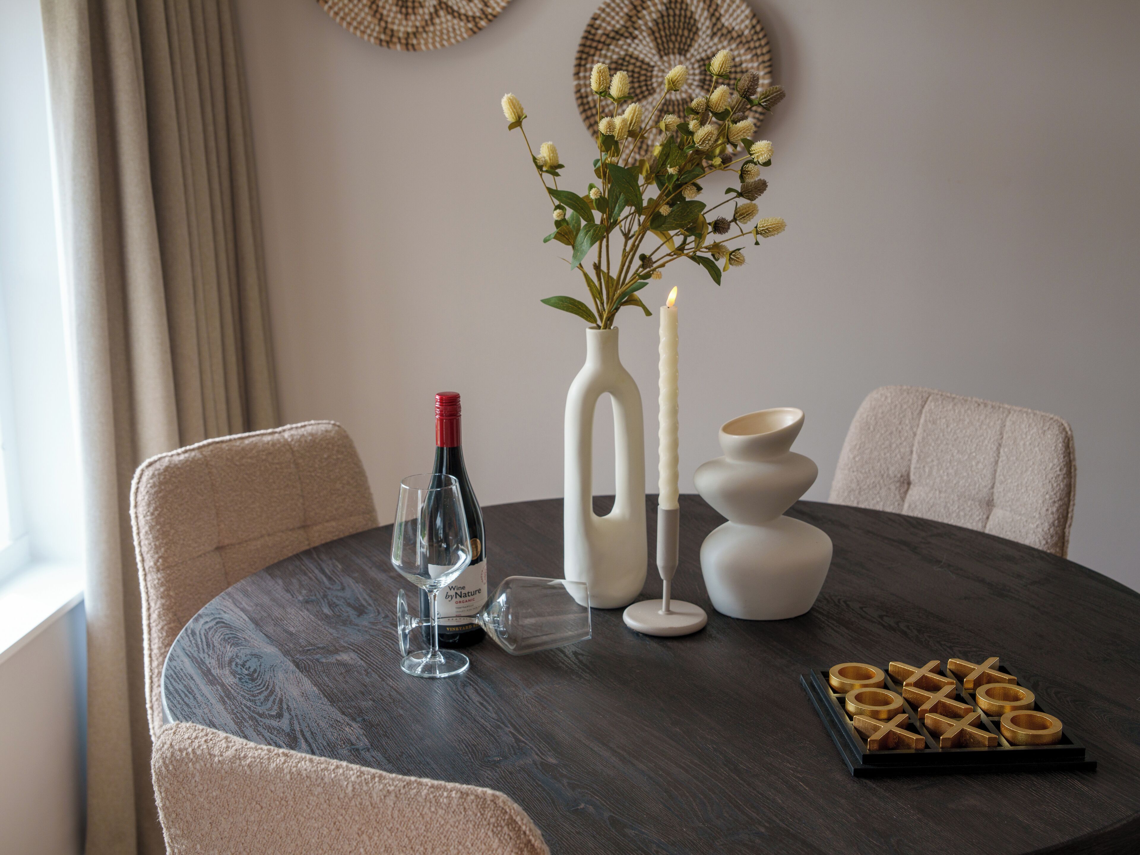 Une table de salle à manger ronde, recouverte d’un vernis en bois foncé, est entourée de chaises beiges à revêtement rembourré. Sur la table, on trouve une bouteille de vin, un verre à vin, un vase blanc avec une plante, une bougie blanche et un plateau contenant des objets ronds et dorés.