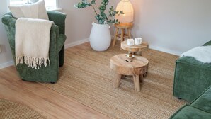 A cozy living room with a green armchair, a white vase with green plants, a wooden side table with candles, and a lamp.