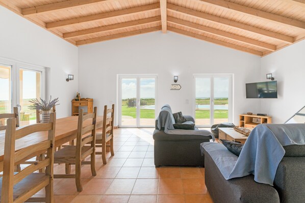 A spacious living area with a high wooden ceiling, a dining table with chairs, a sofa, and a television mounted on the wall.