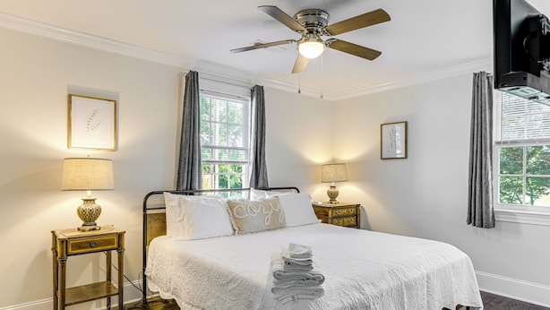 Habitación de hotel con cama, dos mesitas de noche con lámparas, ventilador de techo y ventana con cortinas.