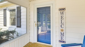 Una puerta azul con un letrero que dice 'WELCOME', un porche con una silla azul y una ventana con contraventanas.