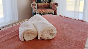 A bed with rolled towels, a patterned armchair, and a window with curtains.