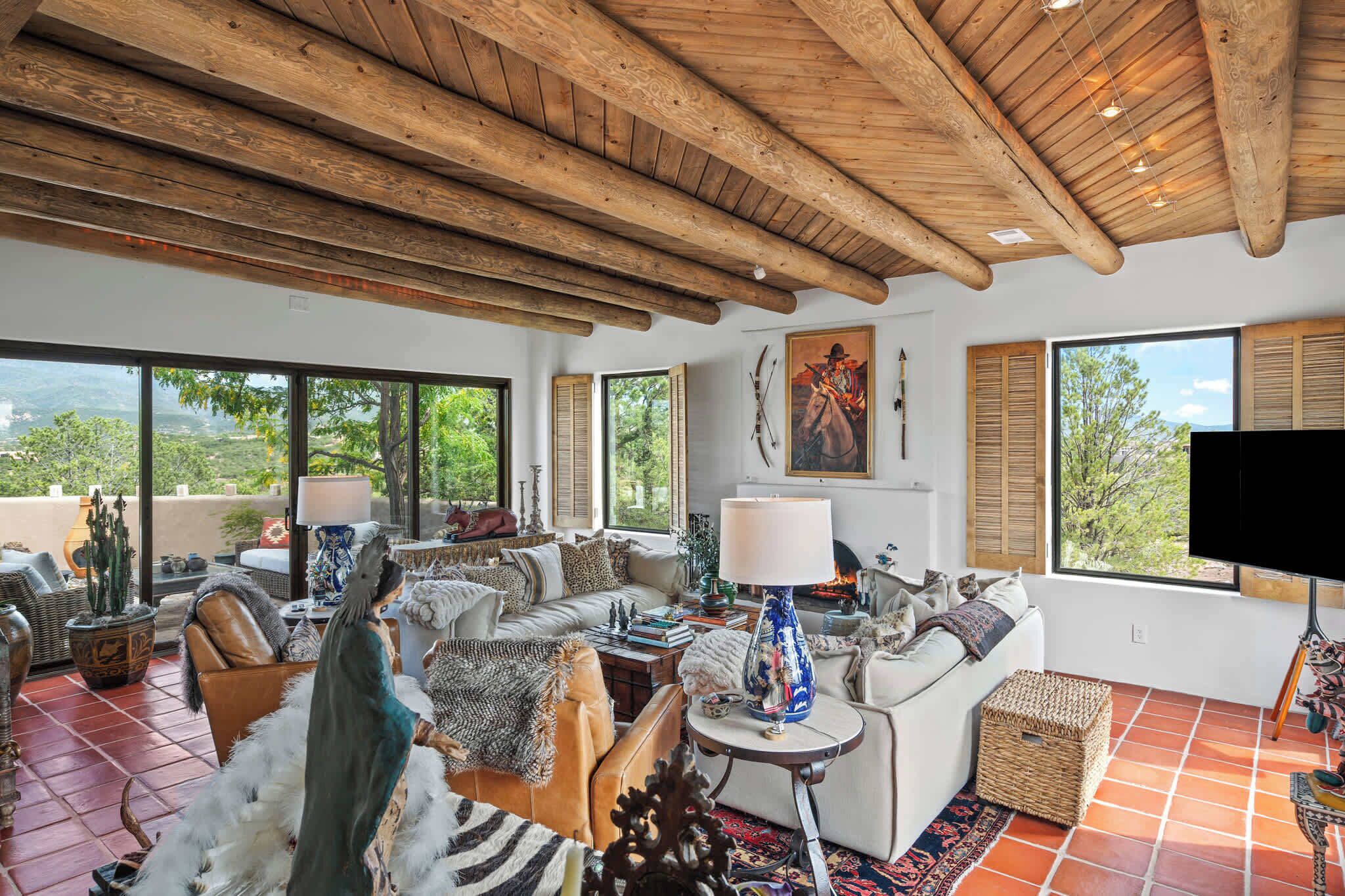 A spacious living room with a high wooden ceiling, large windows offering a view of the outdoors, and a fireplace.