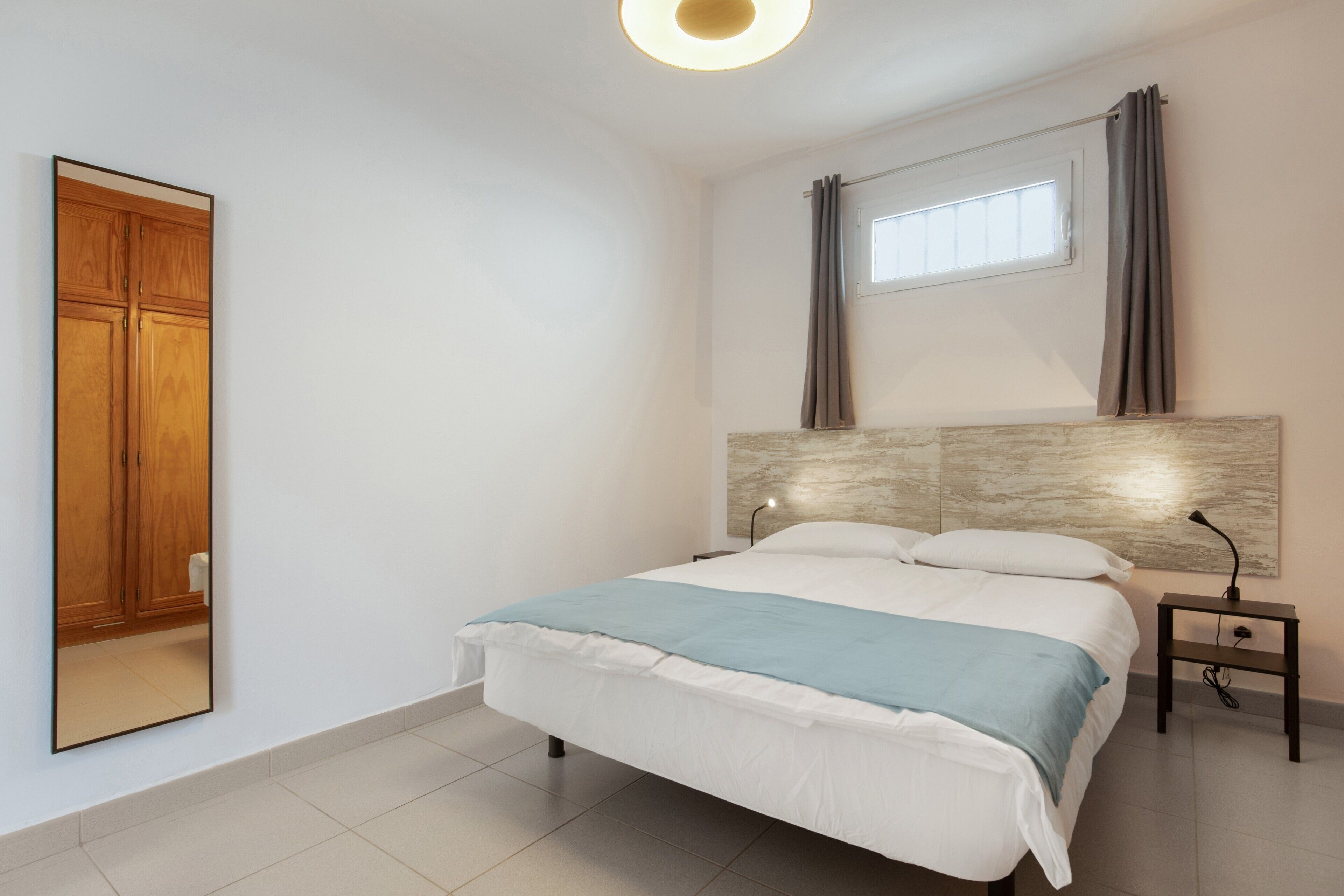 Um quarto com uma cama, uma mesa de cabeceira, um espelho, uma janela com cortinas e um guarda-roupa de madeira.