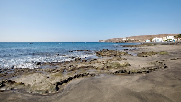 Ein Küstenstrand mit felsigem Gelände, blauem Himmel und weißen Gebäuden im Hintergrund.