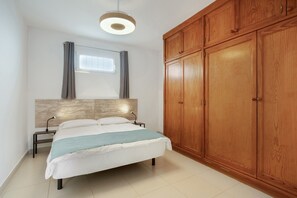 Une chambre à coucher comprenant un lit, une table de chevet, une armoire en bois, un luminaire au plafond et une fenêtre avec des rideaux.