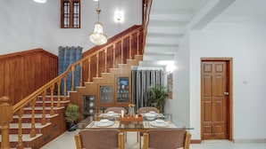 A dining area with a glass table set for four, wooden chairs, a chandelier, a staircase with wooden railings, and a door in the background.