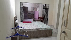 A bedroom with a bed, a small desk, a chair, and a wardrobe. There is a kitchen area visible through an open door. - SAGAR NIWAS (Anekal)