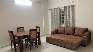 A living room with a brown sofa, a wooden dining table with chairs, and patterned curtains over a window. - SAGAR NIWAS (Anekal)