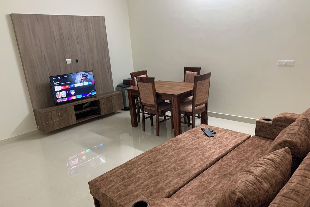 A living room with a flat-screen TV on a wooden stand, a brown sofa, a dining table with chairs, and a small coffee table.